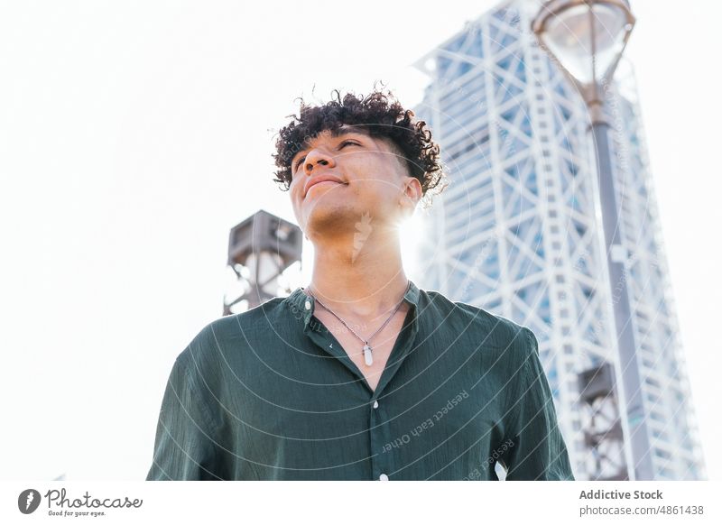 Young ethnic man smiling and looking away on street smile carefree city skyscraper gaze bright summer urban afro modern style guy town personality individuality