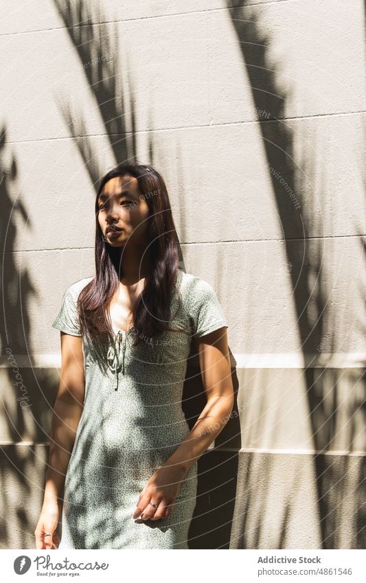 Thoughtful Asian woman near building with shadows wall style sunlight street urban summer charming feminine outfit asian ethnic female city dress lady town