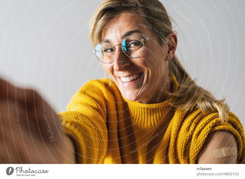 Attractive mature woman smiling after getting a corona vaccine. Woman holding up her shirt sleeve and showing her arm with bandage after receiving vaccination, stretching arm to camera for a selfie.