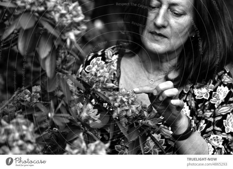 Portrait of an adult woman with dark shoulder-length hair in flowered summer dress, half hidden by a flowering bush. With tactfulness concentrated and gently she touches a blossom