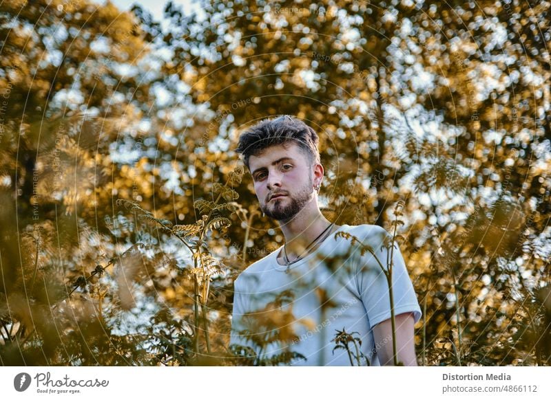 Portrait of a fashionable and mysterious blond young man with blue eyes hidden behind some trees at sunlight thoughtful self love serotonin confident outdoor