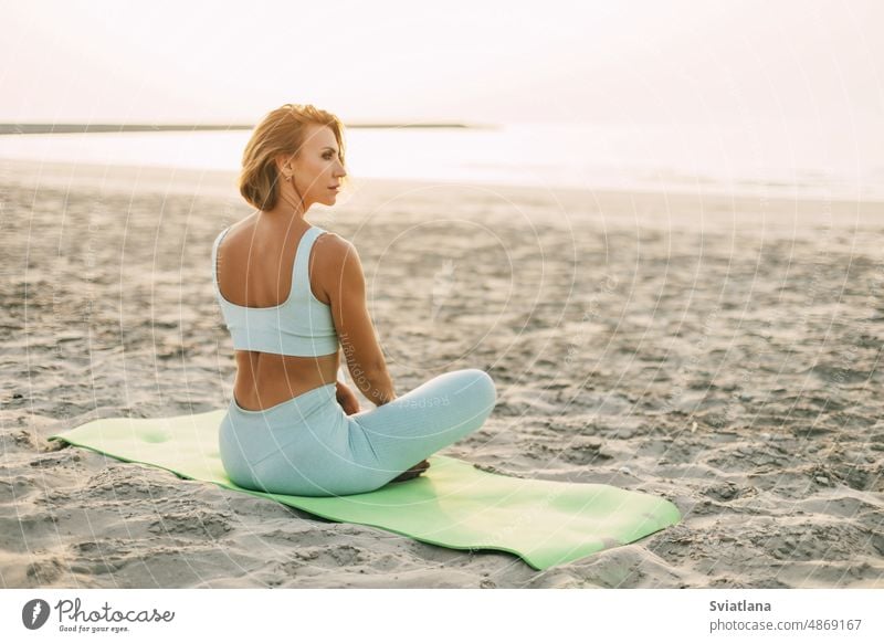 A beautiful girl is sitting on the seashore in the lotus position and resting after yoga or fitness classes. Healthy lifestyle, self-care, activity beach woman