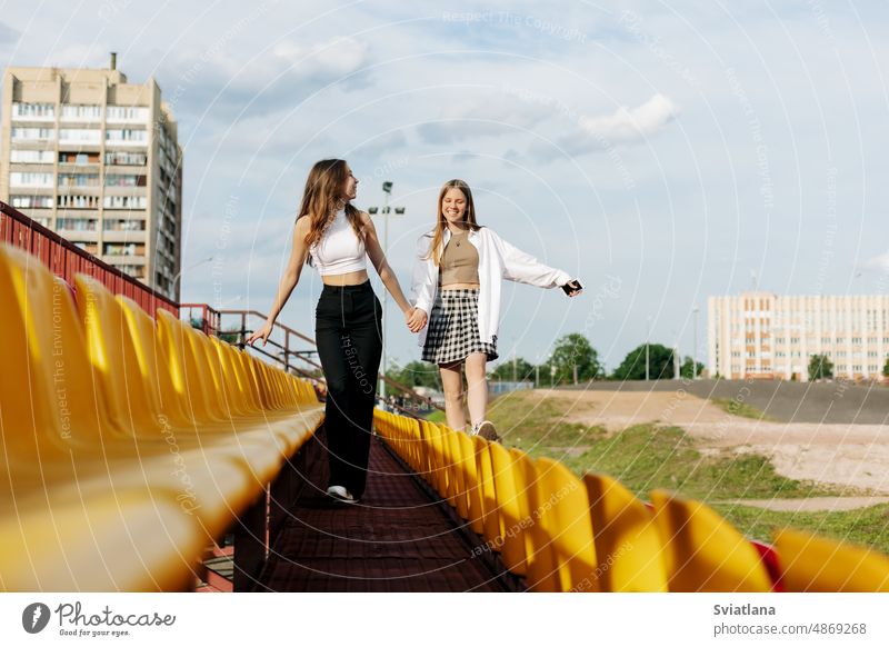 Two teenage girls walk together through the stands of the school stadium, talking, holding hands, best friends return home after training friendship student