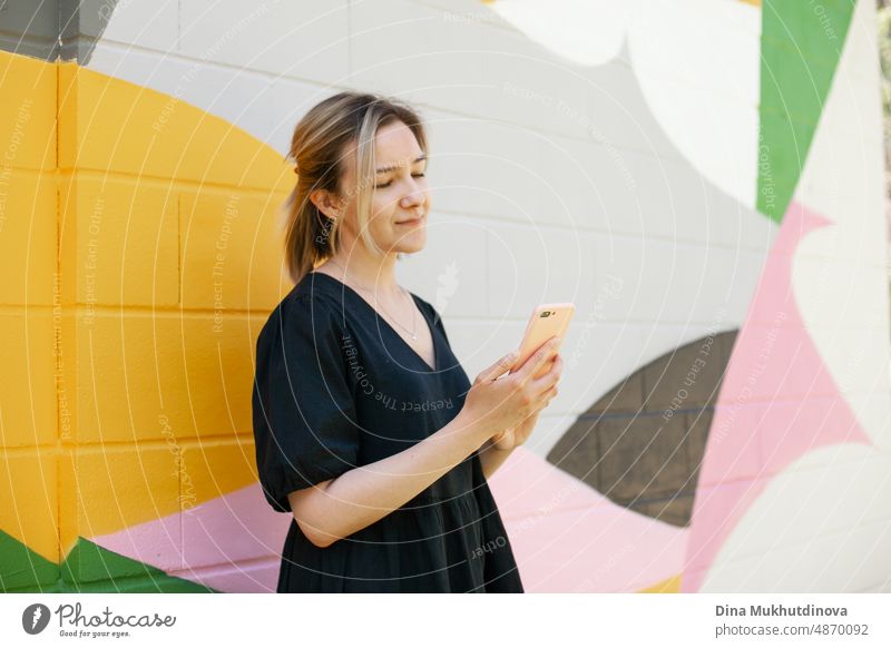 Young woman with cell phone standing near colorful wall taking selfie or video chat on smartphone. Screen time. Lifestyle with technology. Young remote worker employee in modern office space.