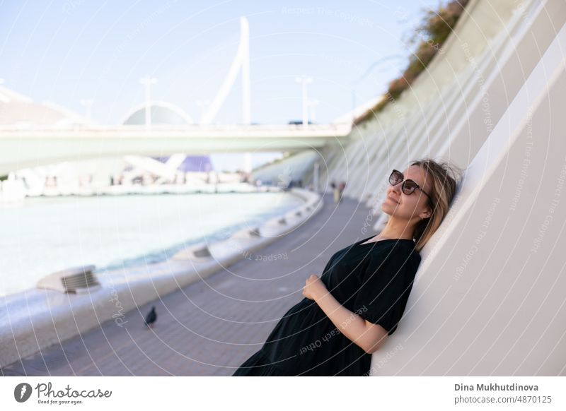 Young female tourist in sunglasses admiring the view of European city on sunny day. Woman sightseeing in summer on vacation in place with modern architecture on background. Heatwave in Europe affecting tourists and travelers.