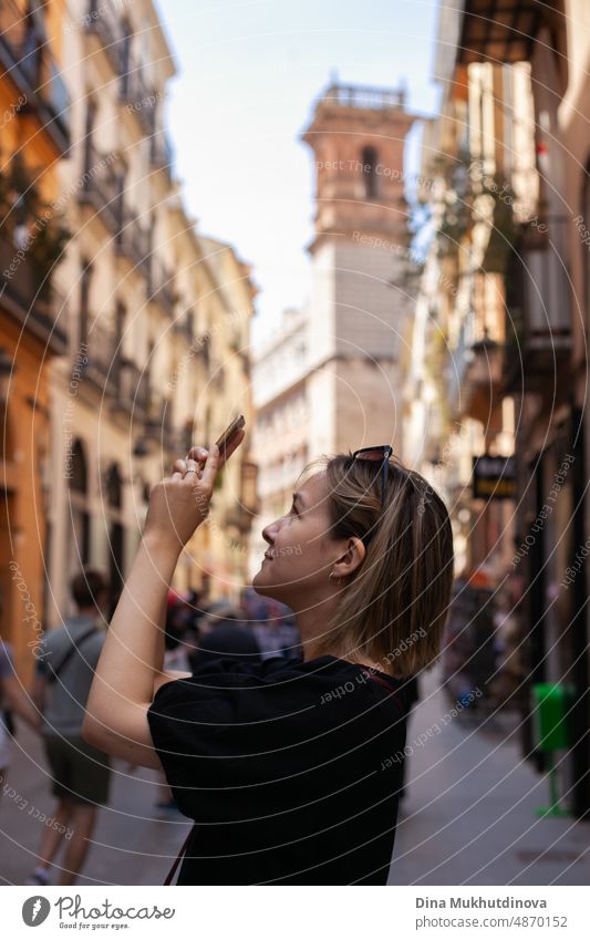 Young female tourist taking pictures with mobile phone in old town admiring the view of European city on sunny day. Woman sightseeing in summer on vacation. Heatwave in Europe affecting tourists and travelers.