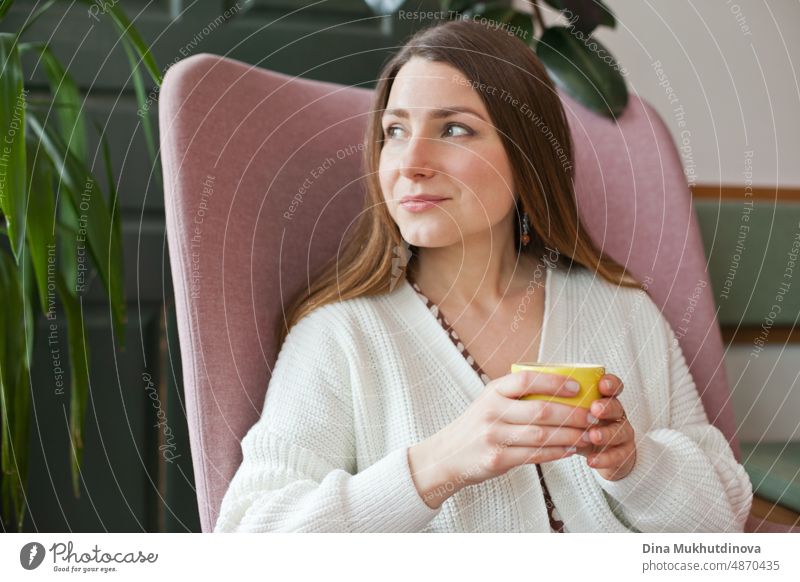 Beautiful woman drinking coffee at cafe sitting in pink chair. Candid millennial lifestyle. Attractive face of an adult woman in her thirties. Looking out the window at restaurant, holding cup of coffee. Coffee break in cafeteria at work in modern office.