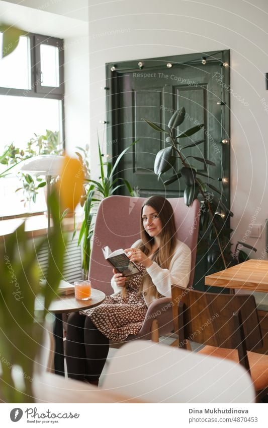 Beautiful woman drinking coffee at cafe sitting in pink chair, reading a book. Student studying at library cafe. Candid millennial lifestyle. Looking out the window at restaurant, holding cup of coffee. Coffee break in cafeteria at work in modern office.