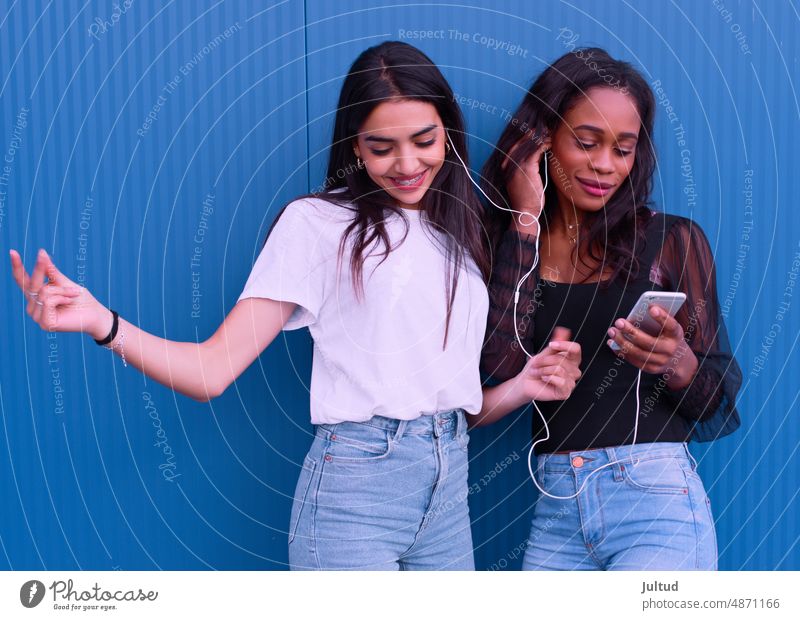 Two friends of different ethnicity listen to music in front of a blue wall. Girl Ethnicity spain Young Woman Twenties Trendy BuildingExterior Girlfriend