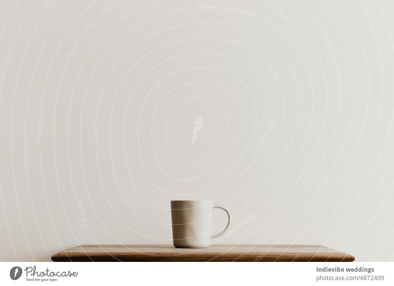 A beige ceramic mug on a brown wooden board in front of a white wall. Hand made coffee cup with copy space. Minimal concept, simple design. tea cafe isolated