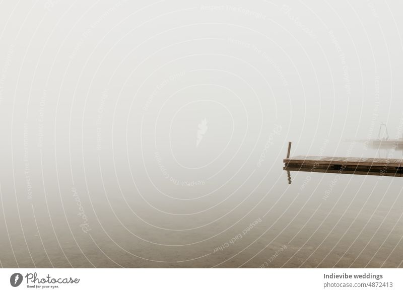 A wooden pier on a lake on a foggy morning. The horizont is not visible due to thick haze. Empty image, copy space. There is no boat by the pier. Lakeside scenery on a cold autumn morning. Nobody.