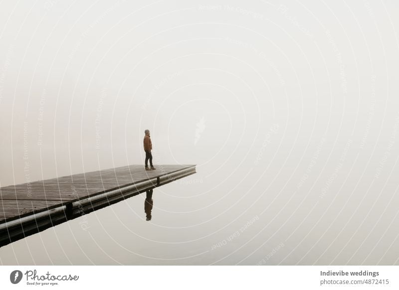 A man is standing on a wooden pier at lake Bohinj in Slovenia. He is facing the distance. The background is foggy. Moody hazy winter landscape. He is thinking, and contemplating. Copy space.