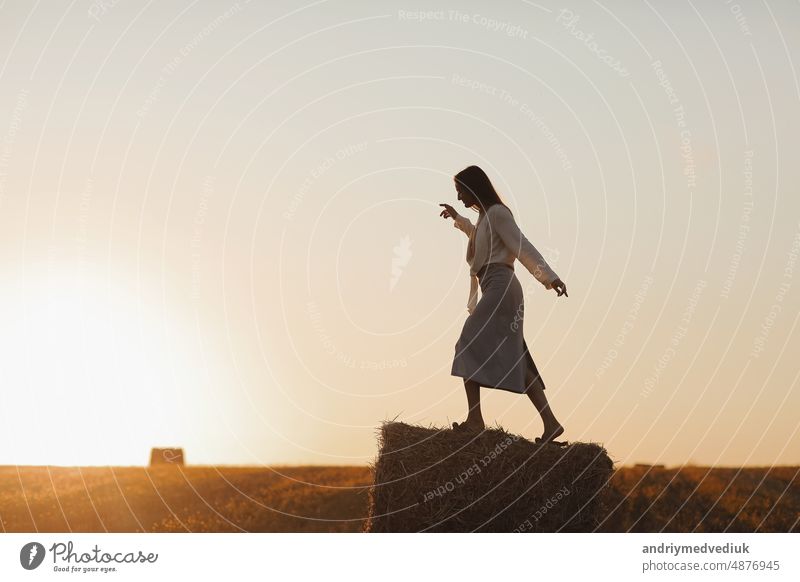 Young woman with long hair is standing and having fun on straw bale in field in summer on sunset. Female portrait in natural rural scene. Environmental eco tourism concept.