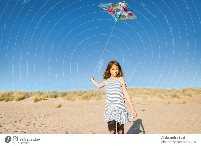 Cute happy little girl in summer dress running with flying kite on empty sandy beach. Beautiful sunny day, blue sky. active activity beautiful carefree cheerful