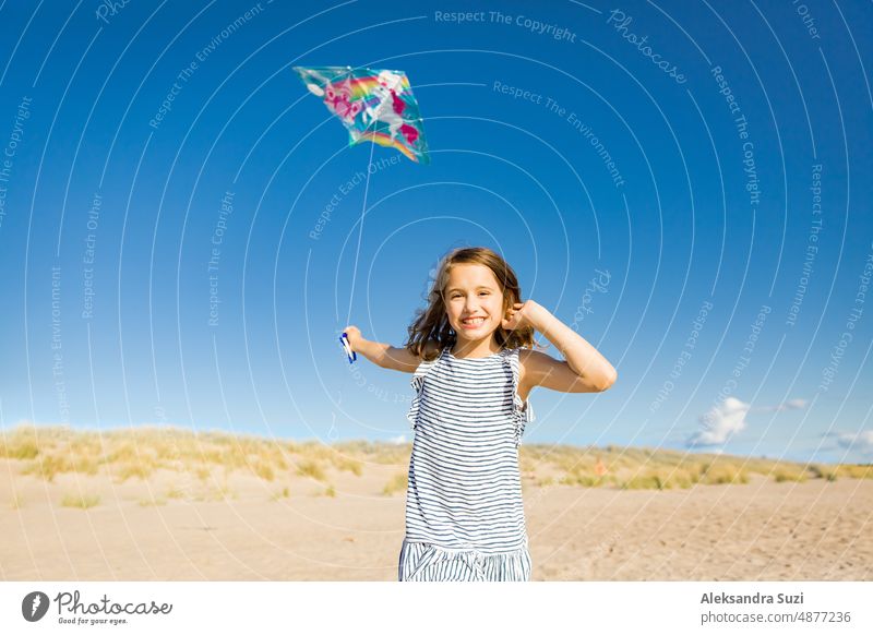 Cute happy little girl in summer dress running with flying kite on empty sandy beach. Beautiful sunny day, blue sky. active activity beautiful carefree cheerful