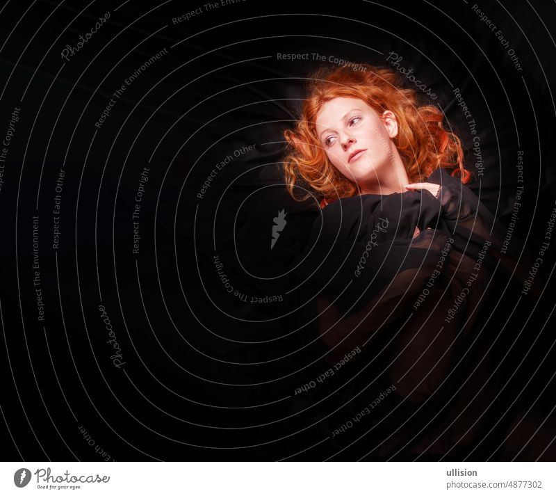 portrait of a beautiful seductive sensual, young sexy redhead, a beautiful ginger woman with long curly red hair with black cloth, lying on black background