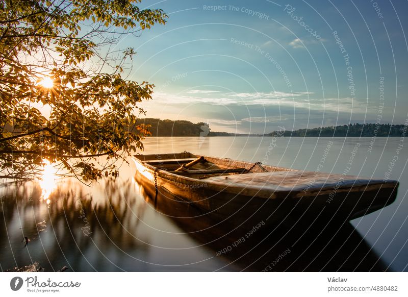 Fairy-tale boat moored on the shore of the Žermanice dam with the setting sun coming through the trees illuminates the breathtaking scenery. Moravian-Silesian region, Czech republic