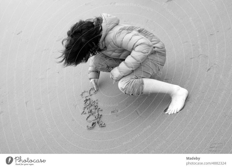 A boy with long hair and a thick down jacket writes something in the sand on the beach in Knokke-Heist on the North Sea near Bruges in West Flanders in Belgium, photographed in classic black and white