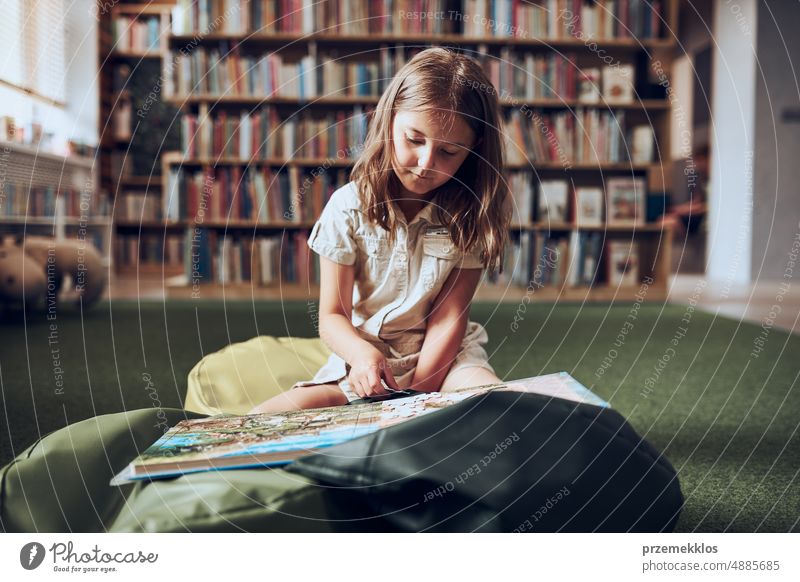 Schoolgirl doing puzzle and reading book in school library. Primary school pupil is involved in book with jigsaw. Benefits of everyday reading. Child curiosity