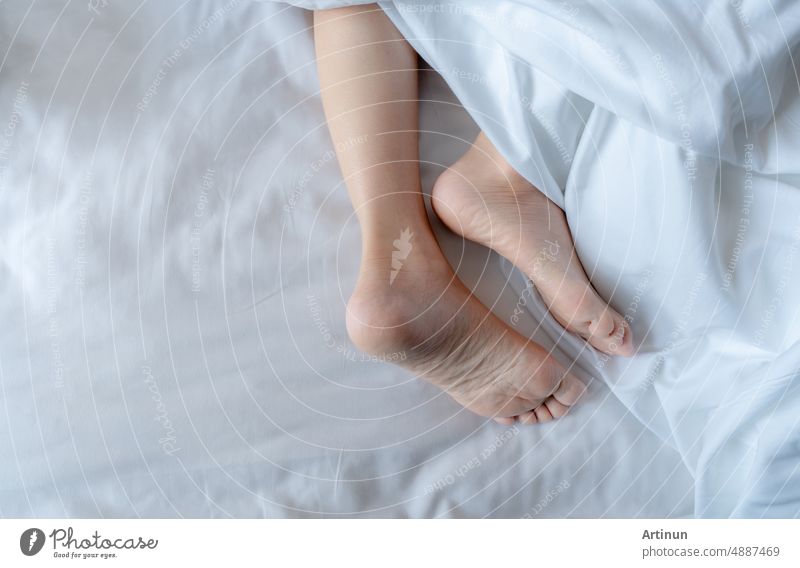 Woman barefoot on bed under white linen blanket in hotel or home bedroom. Healthy sleep and relaxation concept. Lazy Sunday morning. Bare feet of woman chilling sleep on white comfort bed and duvet.