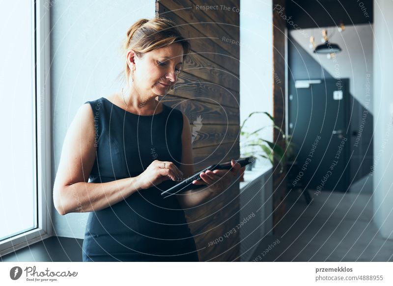 Businesswoman working on tablet in office. Mature woman using touch pad computer standing by window in modern interior. Manager focused on work holding digital device. Using technology