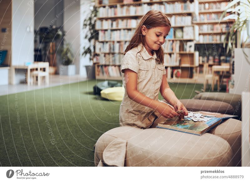 Schoolgirl doing puzzle and reading book in school library. Primary school pupil is involved in book with jigsaw. Benefits of everyday reading. Child curiosity