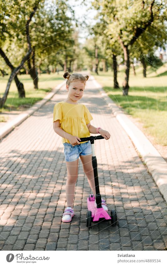 An active little girl rides a scooter on a path in an outdoor park on a summer day. Seasonal children's active sport. Healthy lifestyle in childhood outdoors