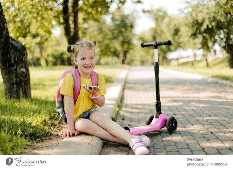 A charming little girl with a phone in her hand, with a backpack and a scooter is sitting on the sidewalk after school. Back to school, after school, school age