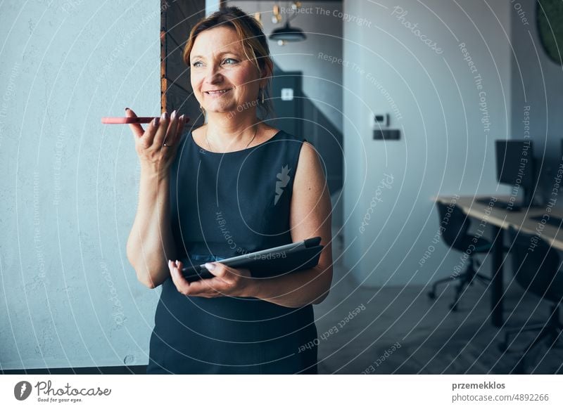 Businesswoman having business call working on tablet in office. Mature busy woman using touch pad computer standing by window in modern interior smartphone talk