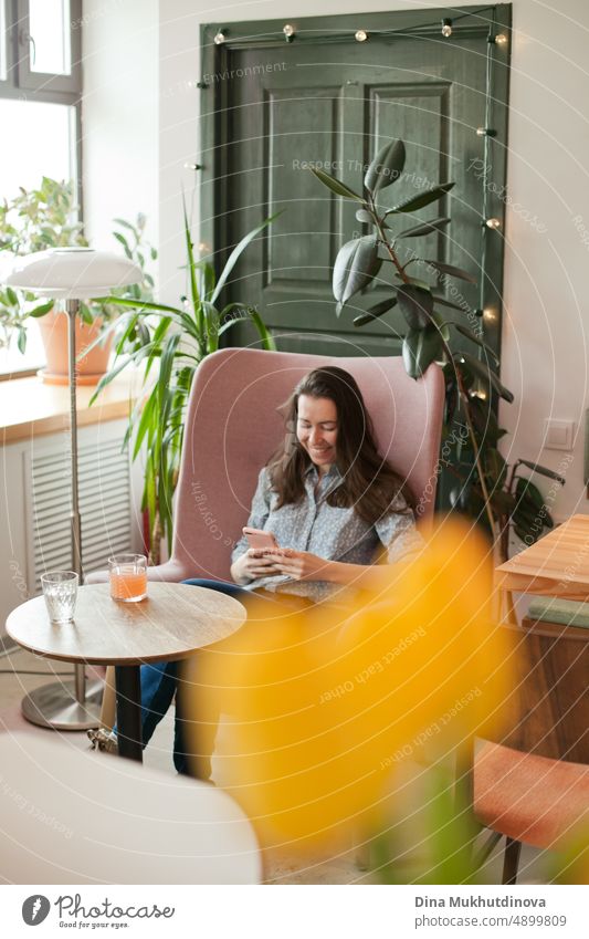 Young millennial woman using mobile phone sitting in cafe chatting with friends. Real person in interior of restaurant, smiling happily.  Using technology, texting and drinking juice. Hipster drinking juice healthy lifestyle.