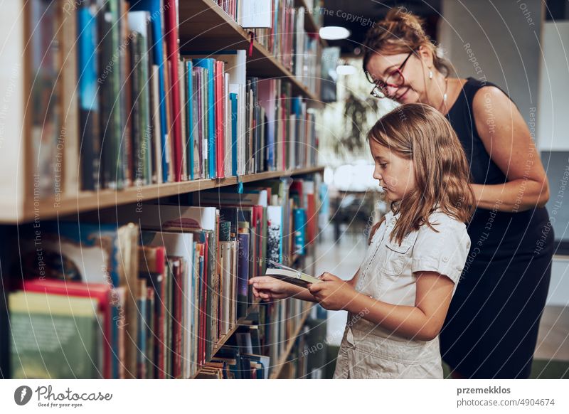 Teacher helping to choose book her schoolgirl in school library. Smart girl selecting literature for reading. Books on shelves in bookstore. School education. Benefits of everyday reading