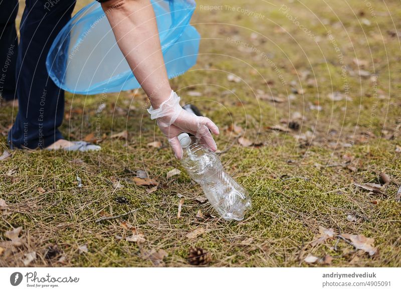 A woman hands in gloves collects and puts used plastic bottle into a blue trash bag. A volunteer cleans up the park on a sunny bright day. Clearing, pollution, ecology and plastic concept