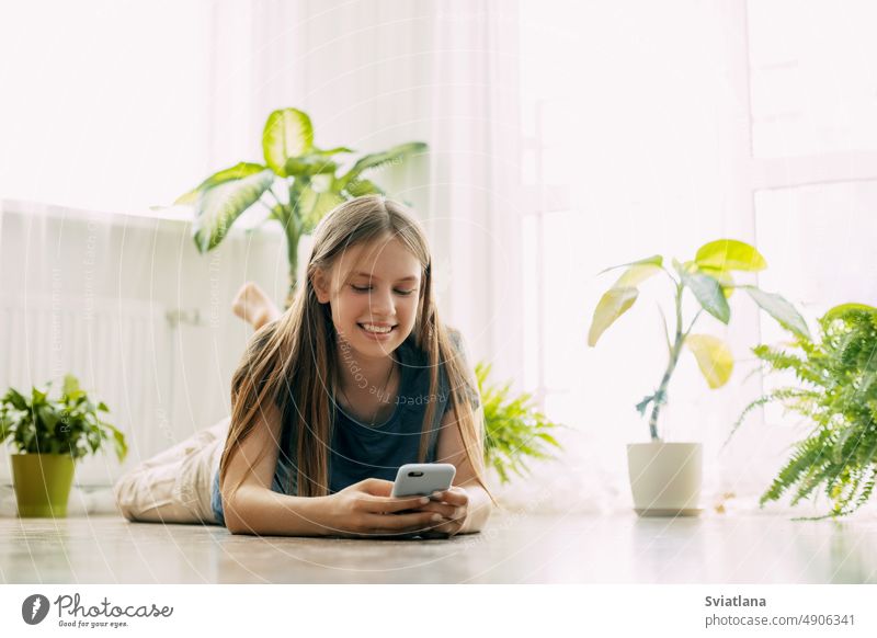 A happy teenager is texting on a mobile phone, lying on the floor in the living room. Modern technologies, online communication student girl technology message