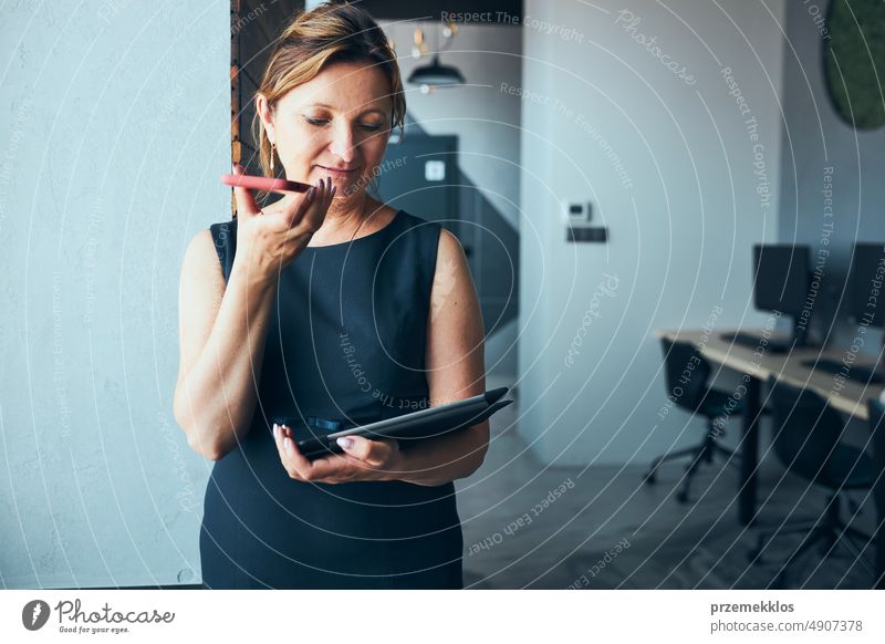 Businesswoman having business call working on tablet in office. Mature busy woman using touch pad computer standing by window in modern interior smartphone talk