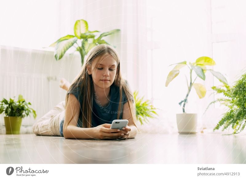 A happy teenager is texting on a mobile phone, lying on the floor in the living room. Modern technologies, online communication student girl technology message