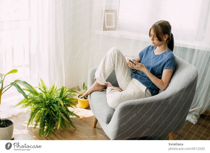 A young girl is sitting in a chair and exchanging messages online on her phone. Home recreation, modern lifestyle student messaging using communication message