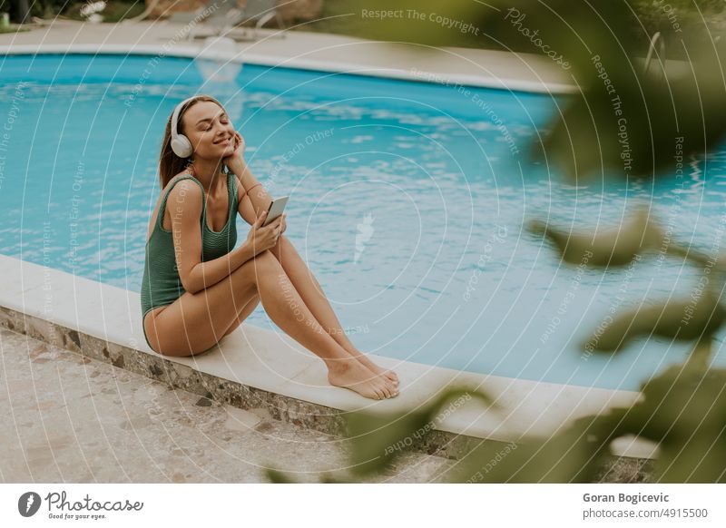 Young woman sitting by the swimming pool and listen music from mobile phone with headphones in the house backyard bikini caucasian day enjoying exterior female