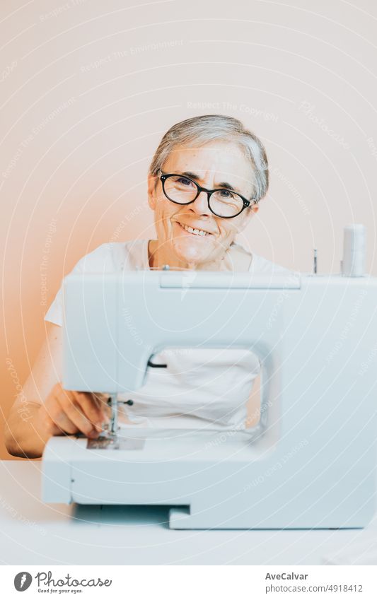 Old woman tailor working on sewing machine smiling happy to camera while sew. Re-use old fabrics for new clothes, sustainability. Old people new hobby at home, seniors mental activity and job