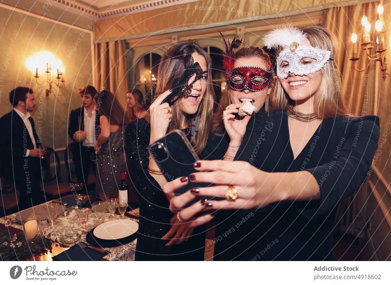 Positive women in carnival masks taking selfie in restaurant self portrait dinner masquerade friend having fun social media occasion bonding pastime enjoy