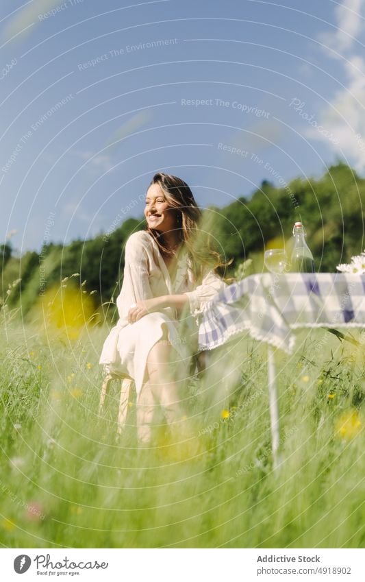 Positive woman sitting at table in field picnic countryside summer recreation nature grass pastime rest feminine smile beverage female dress optimist content