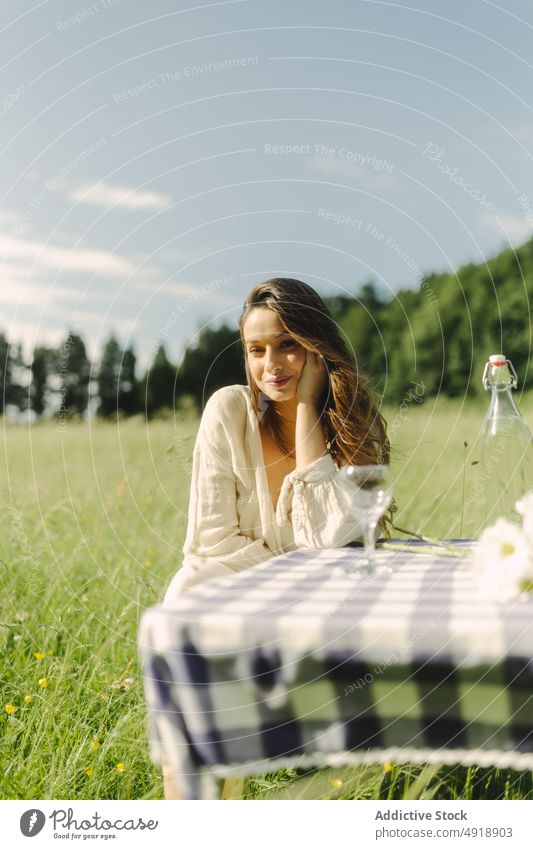 Positive woman sitting at table in field picnic countryside summer recreation nature grass pastime rest feminine smile beverage female dress optimist content