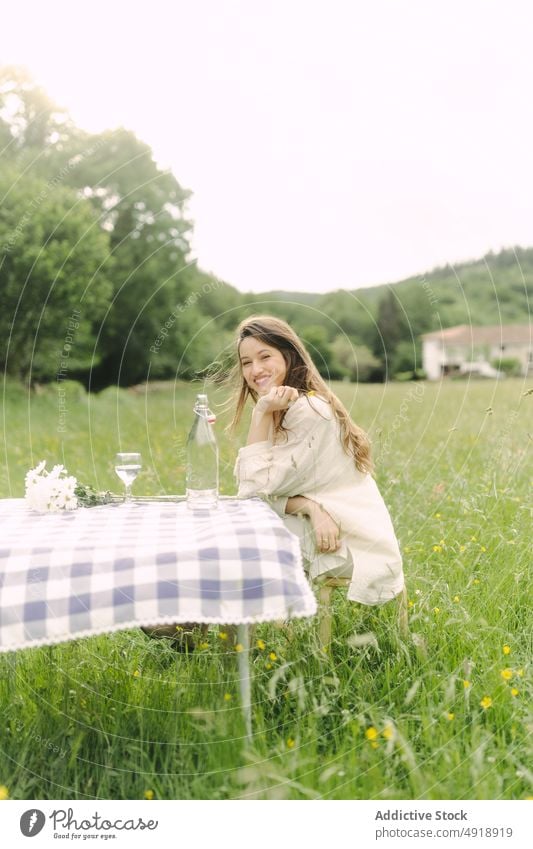 Positive woman sitting at table in field picnic countryside summer recreation nature grass pastime rest feminine smile beverage female dress optimist content