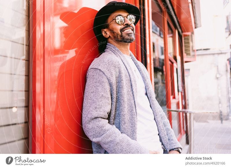 Cheerful Hispanic man leaning on wall street smile building happy street style urban rest male adult hispanic ethnic trendy cheerful sunglasses hat coat