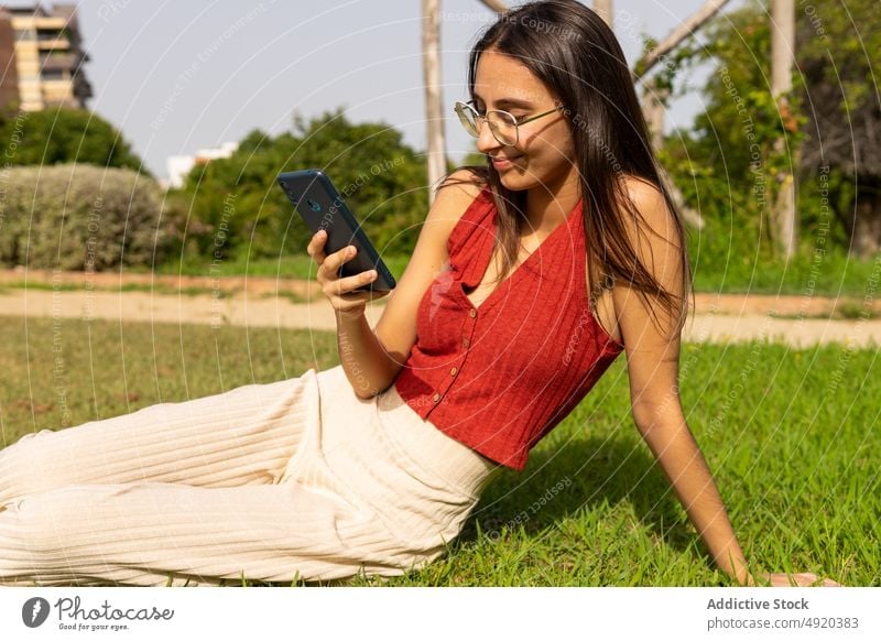 Smiling woman browsing smartphone on lawn rest text message park online social media chat app chill free time scroll positive joy smile cheerful student city