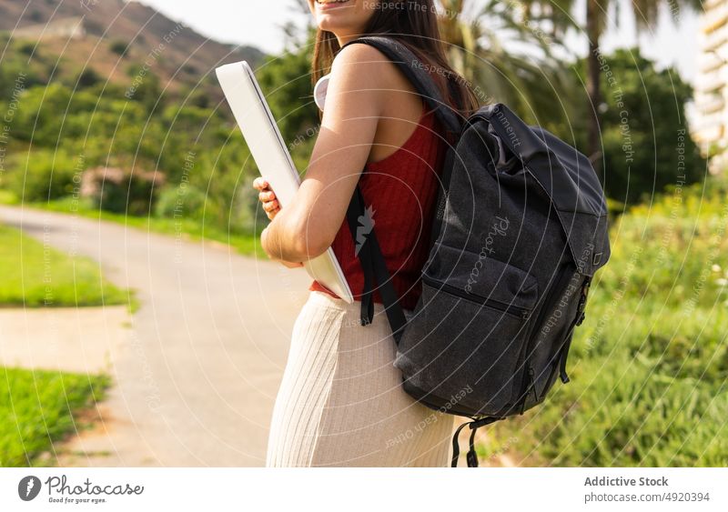 Faceless woman standing in park with a closed computer in his arms summer crop laptop faceless city backpack anonymous female cheerful tree headphones young