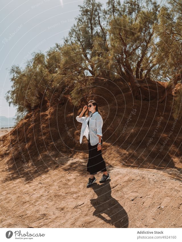 Stylish woman standing near hill with green trees on sunny day desert tourist style calm sand nature trip plant idyllic harmony female young long hair dark hair