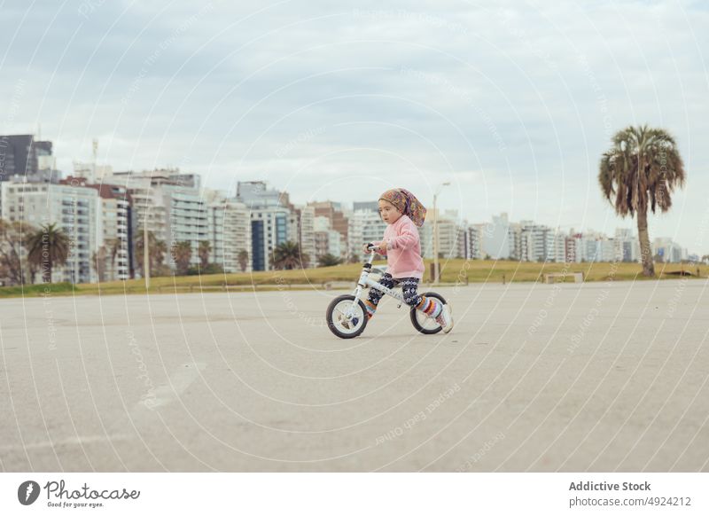 Girl riding bicycle on street girl ride balance urban road weekend cloudy sky apartment building gray way move child kid casual preschooler little daytime