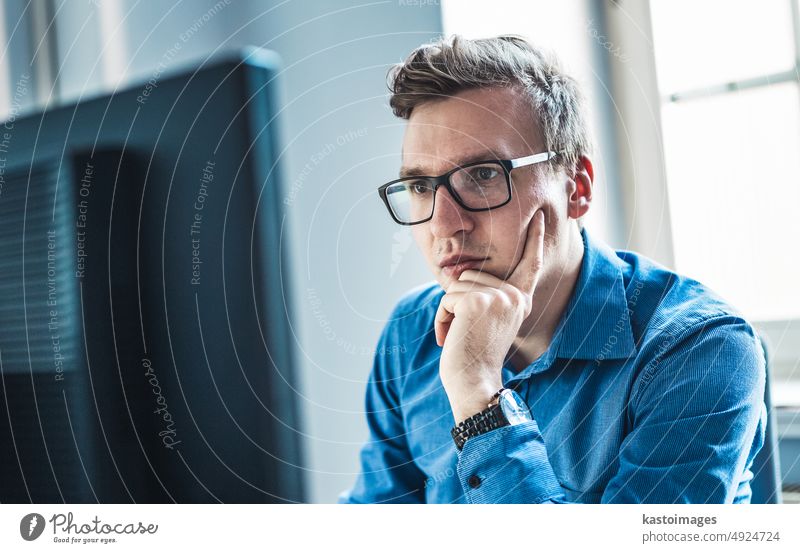 Handsome Young Businessman Wearing Eyeglasses Sitting at his Table Inside the Office, Looking at the Report on his Computer Screen businessman monitor portrait