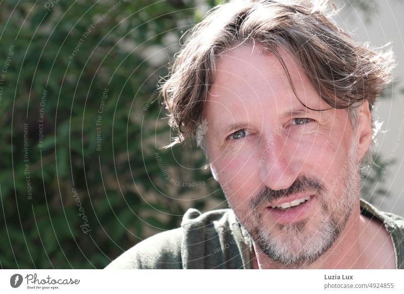 Handsome middle-aged man looking amused at camera, around it a lot of greenery Grinning Disheveled Curl Designer stubble Facial hair Adults kind Smiling