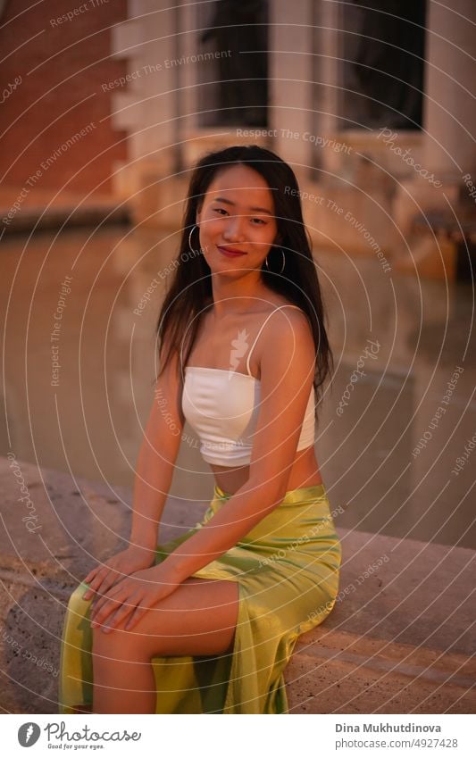 Beautiful young Asian woman sitting near fountain in European historic town at night with streetlights on. Female tourist from China traveling in Europe. Fashion and style concept.